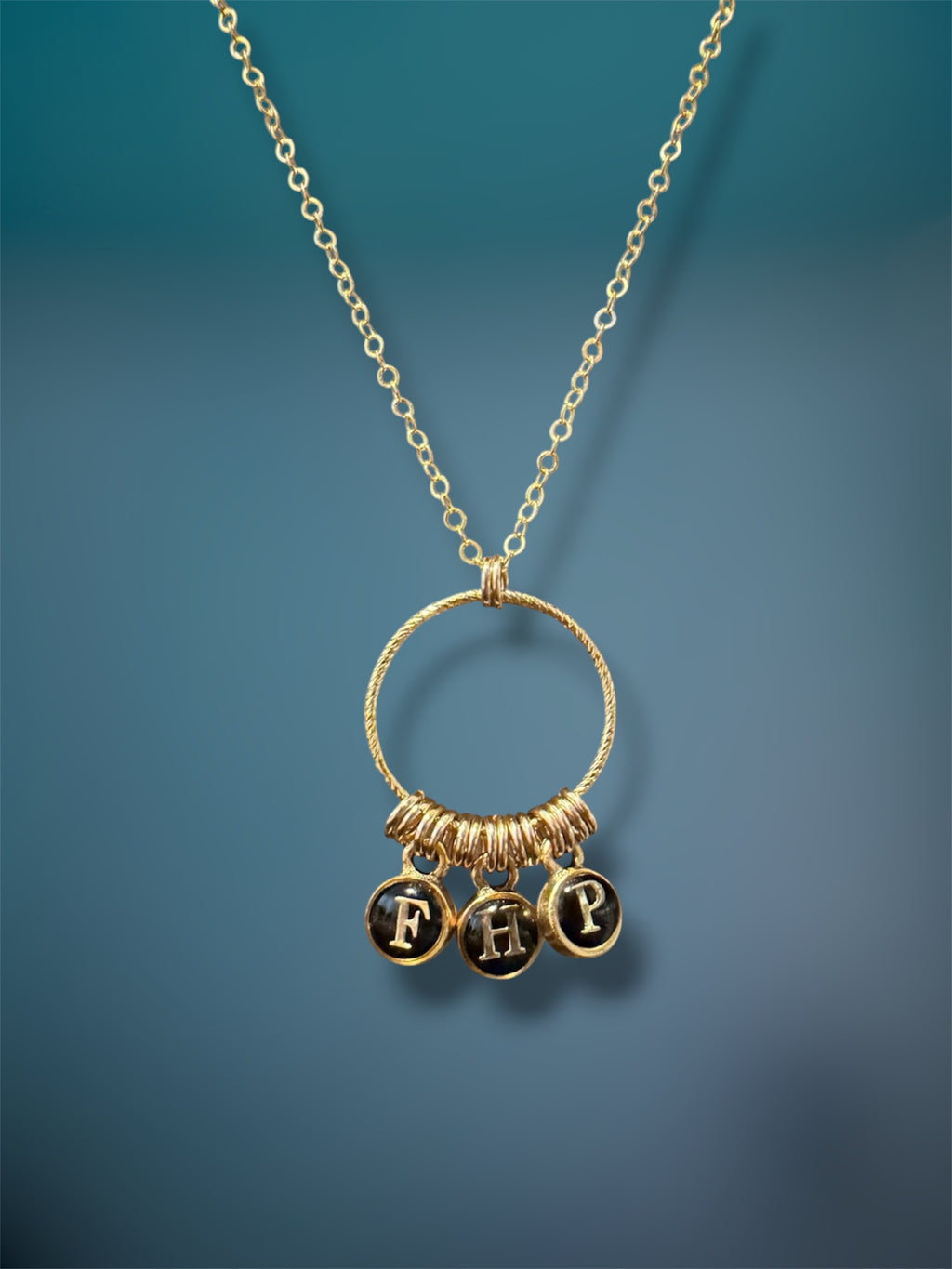 Gold-filled initial charm necklace with three black typewriter-style letter charms F, H, and P on a textured gold ring, teal background