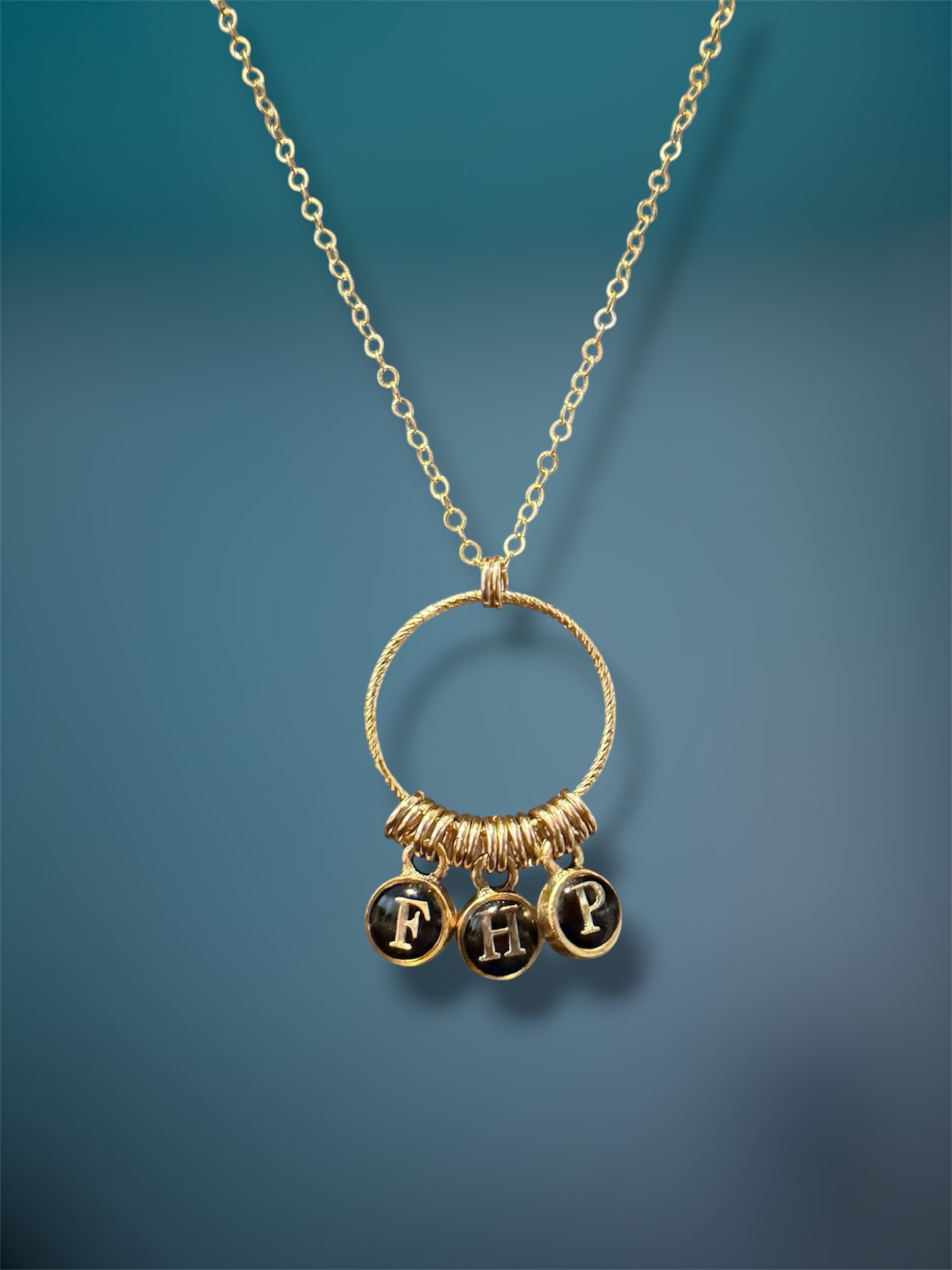 Gold-filled initial charm necklace with three black typewriter-style letter charms F, H, and P on a textured gold ring, teal background